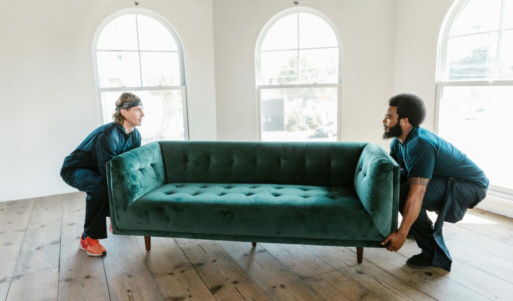 Two men are lifting a green sofa in a bright room with wooden floors and arched windows, rearranging furniture together.