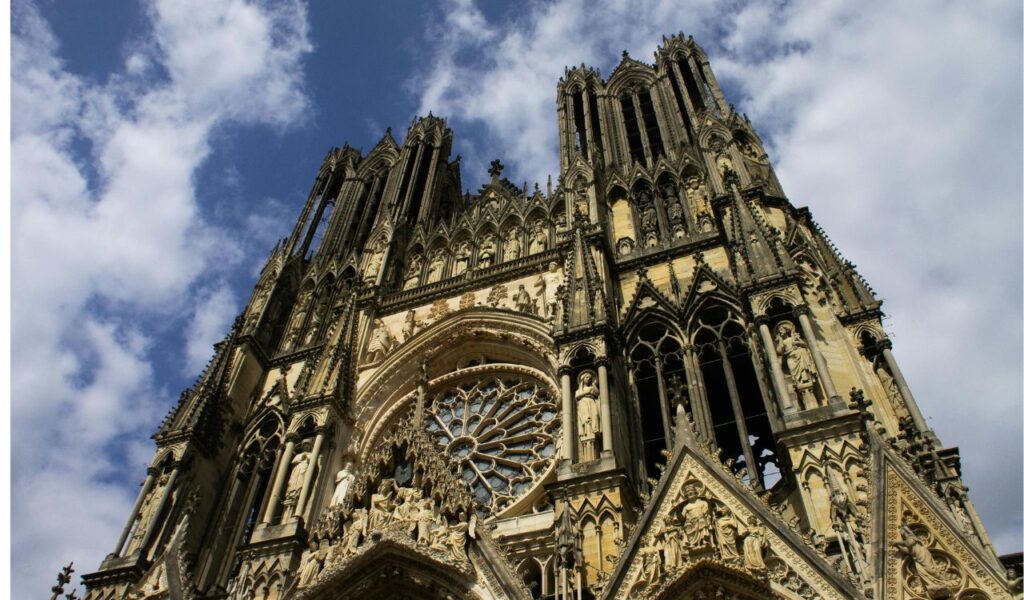 Low-angle view of Reims Cathedral in France, showcasing its intricate Gothic architecture, ornate sculptures, and rose window under a partly cloudy sky.