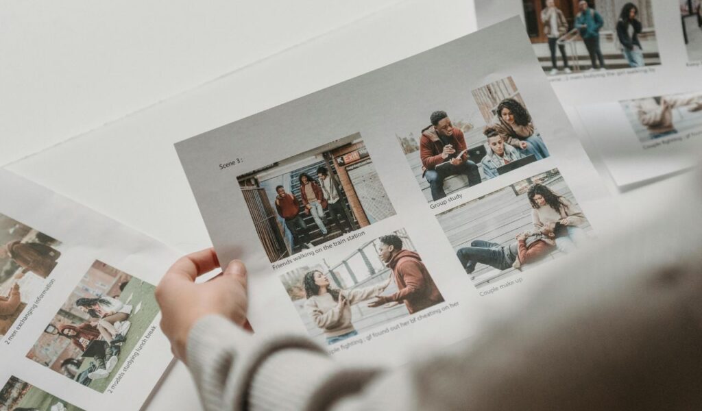 A person holding a printed sheet of photo collages showing friends walking, studying together, and a couple reconciling, with other similar printed sheets spread out on the table.