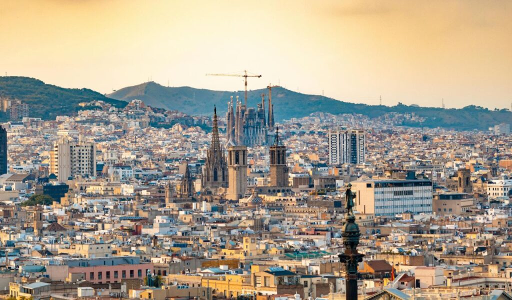 A panoramic view of Barcelona, Spain, with the towering spires of the Sagrada Familia basilica under construction, set against hills and a warm evening sky.