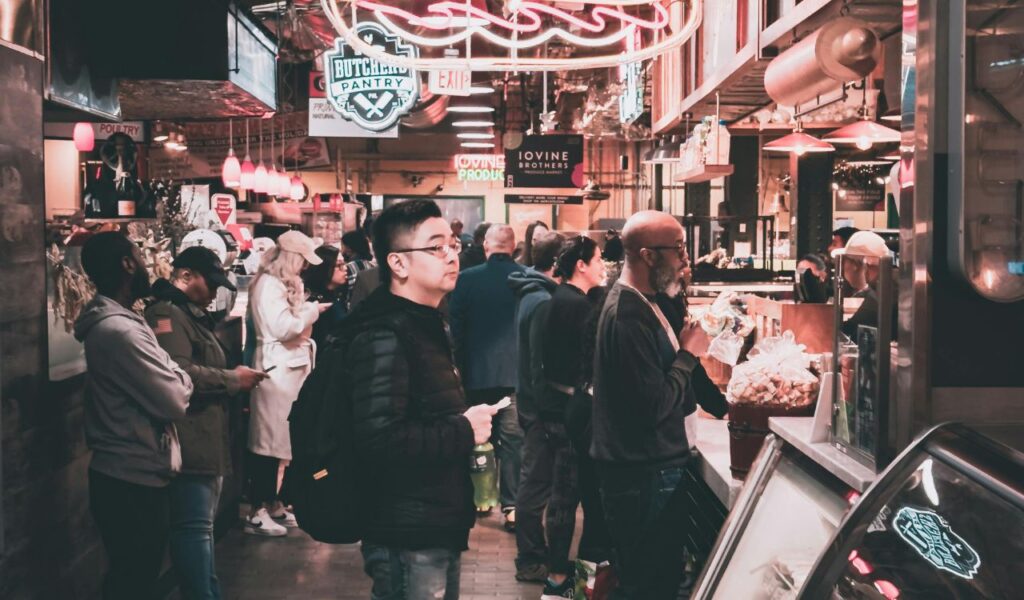 People wait in line inside Savannah’s bustling City Market, surrounded by neon signs, food counters, and displays of local goods.