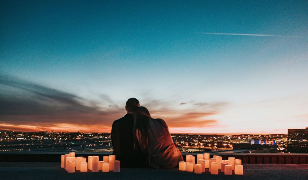 A couple sitting close together surrounded by glowing candles, watching the sunset over a city skyline.
