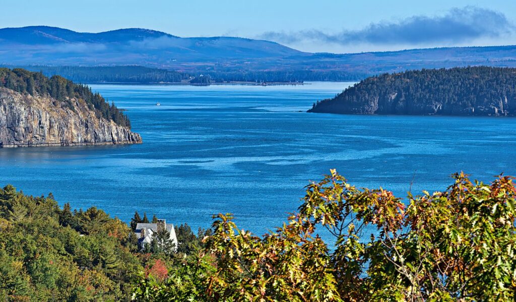 A panoramic view of the Schoodic Peninsula in Acadia National Park shows rugged cliffs, forested shores, and deep blue waters under a clear sky.