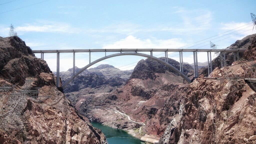 A wide view of the Mike O'Callaghan–Pat Tillman Memorial Bridge spanning over the rocky canyon near the Hoover Dam with blue sky and power lines in the background.