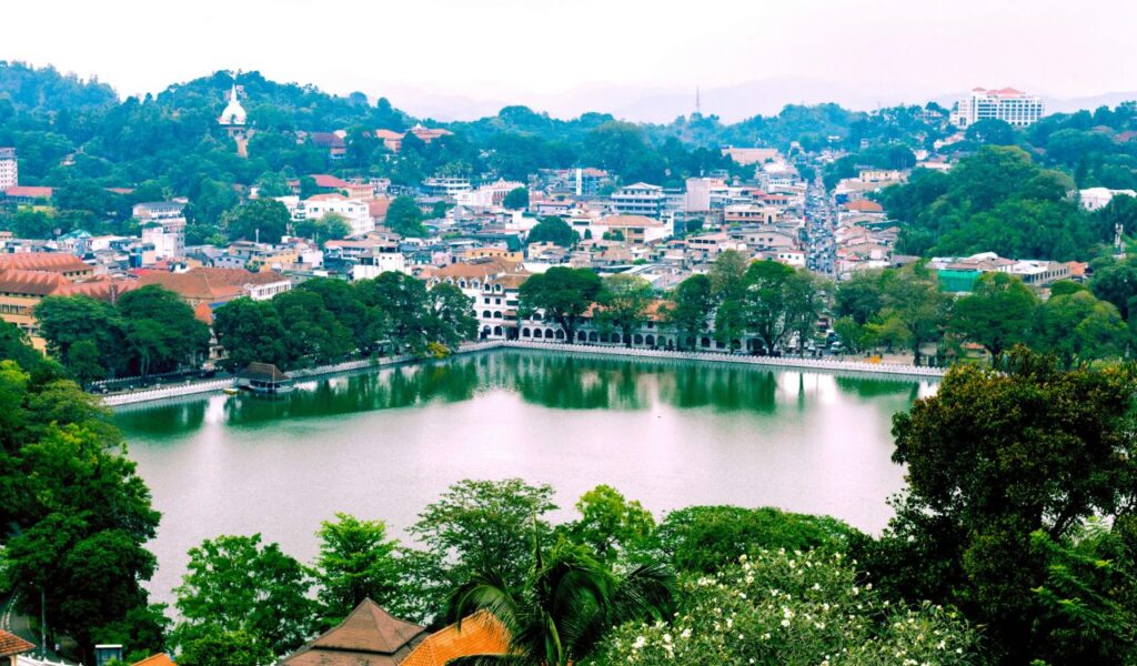 A scenic view of Kandy, Sri Lanka, featuring the peaceful Kandy Lake surrounded by lush greenery, historic buildings, and a hillside dotted with temples and houses.