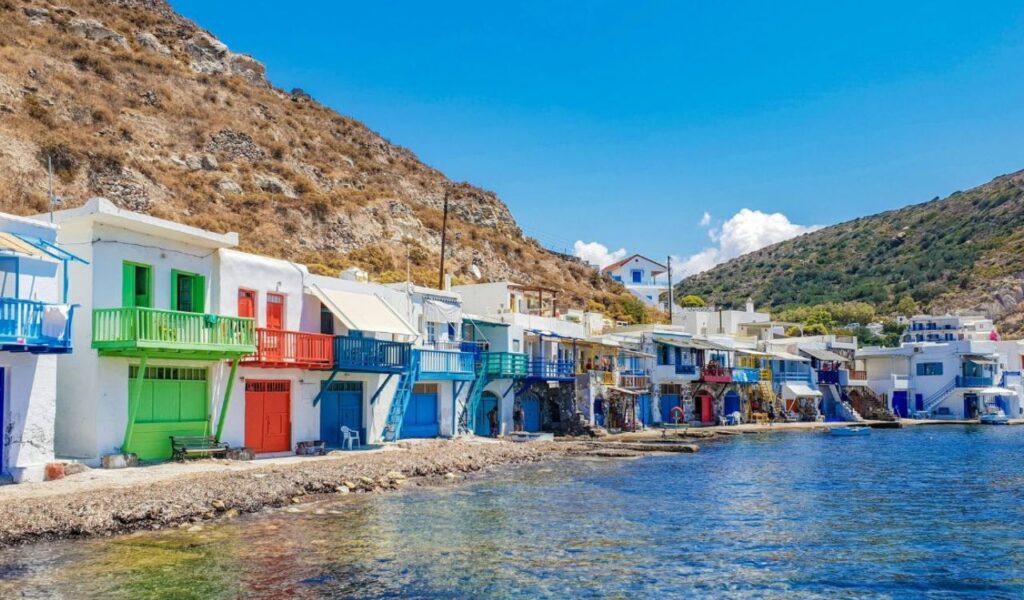 Colorful waterfront houses with bright balconies line the shore of a quiet bay on a Greek island, set against rocky hills under a clear blue sky.
