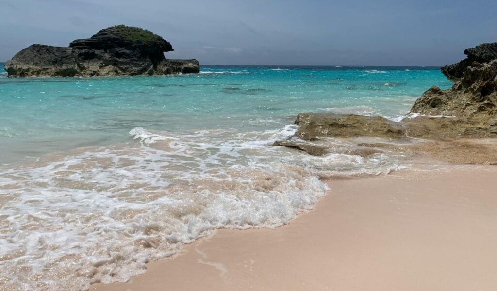 A serene beach with turquoise waves gently rolling onto the soft sandy shore, bordered by rocky outcrops and a distant rock formation.