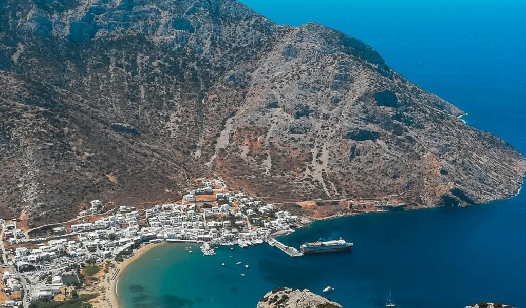 Aerial view of Sifnos, Greece, showing whitewashed houses clustered around a turquoise bay, backed by rugged, rocky hills.
