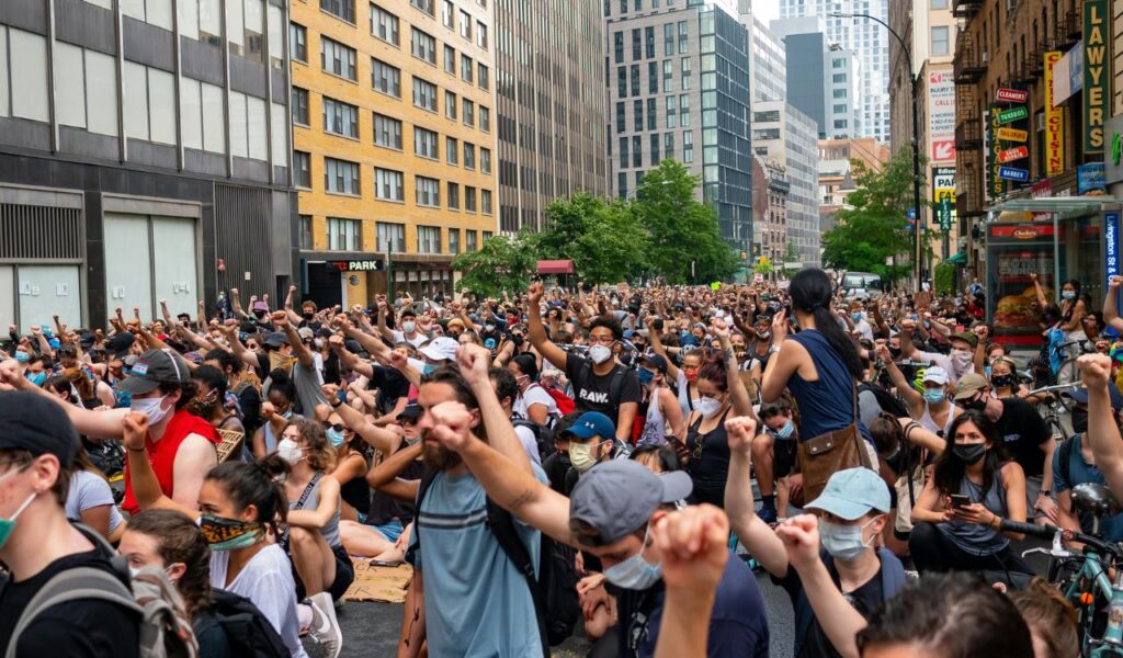 A large crowd of masked protesters raising their fists during a demonstration in a city street surrounded by tall buildings.