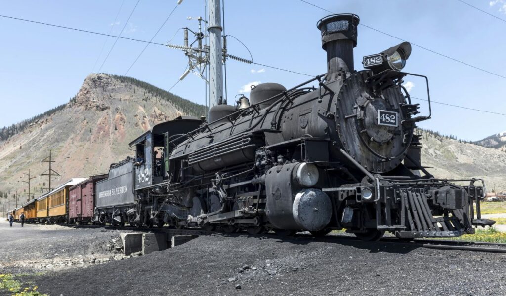 Historic black steam locomotive with attached train cars at a station in Silverton, Colorado, with mountains in the background.