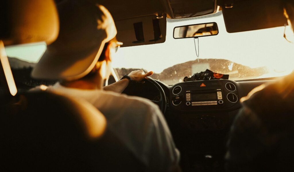A person drives a car at sunset, with warm sunlight streaming through the windshield, creating a relaxed road trip atmosphere.