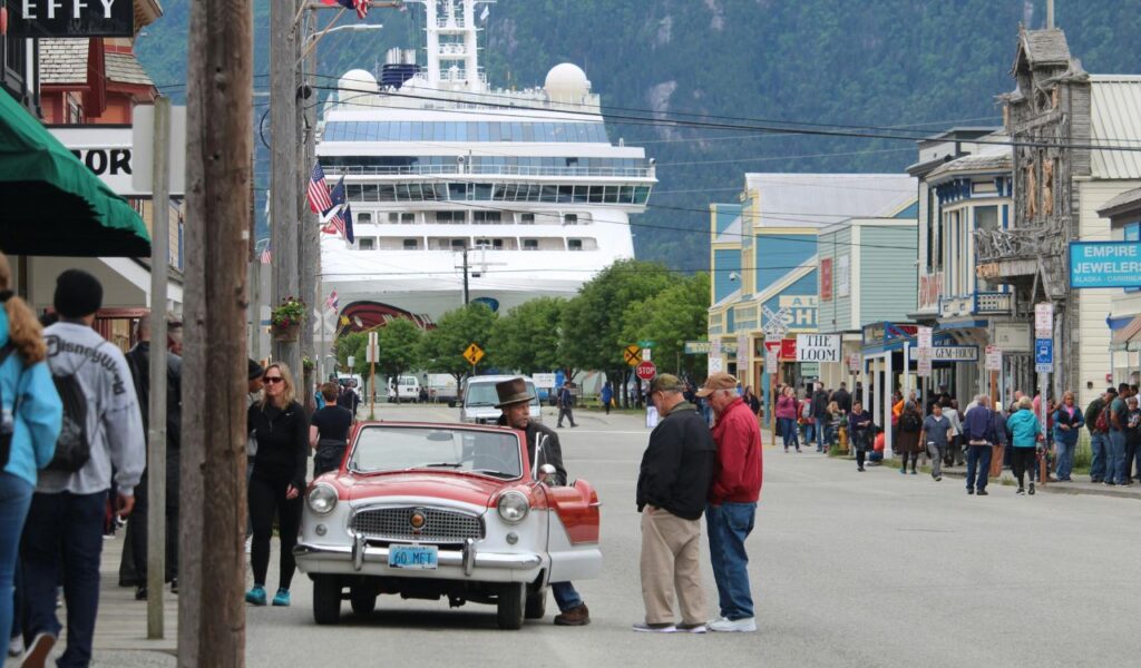 Street scene in Skagway, Alaska, with people walking along shops, a vintage red and white car in the foreground, and a large cruise ship docked at the end of the street.