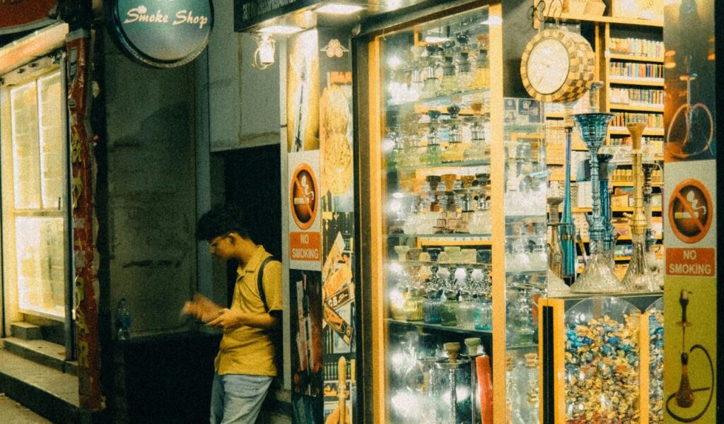 A young man stands outside a brightly lit smoke shop at night, looking at his phone. The shop displays glassware, hookahs, and "No Smoking" signs.