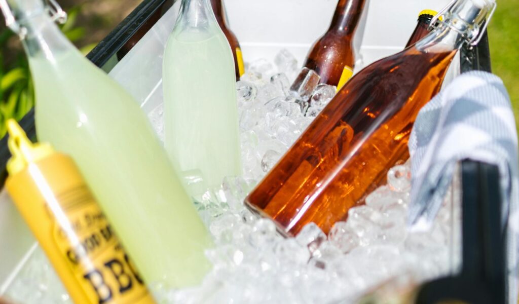 Bottles of drinks and a mustard container sit in a cooler filled with ice, with a towel draped over the side, ready for a picnic or trip.