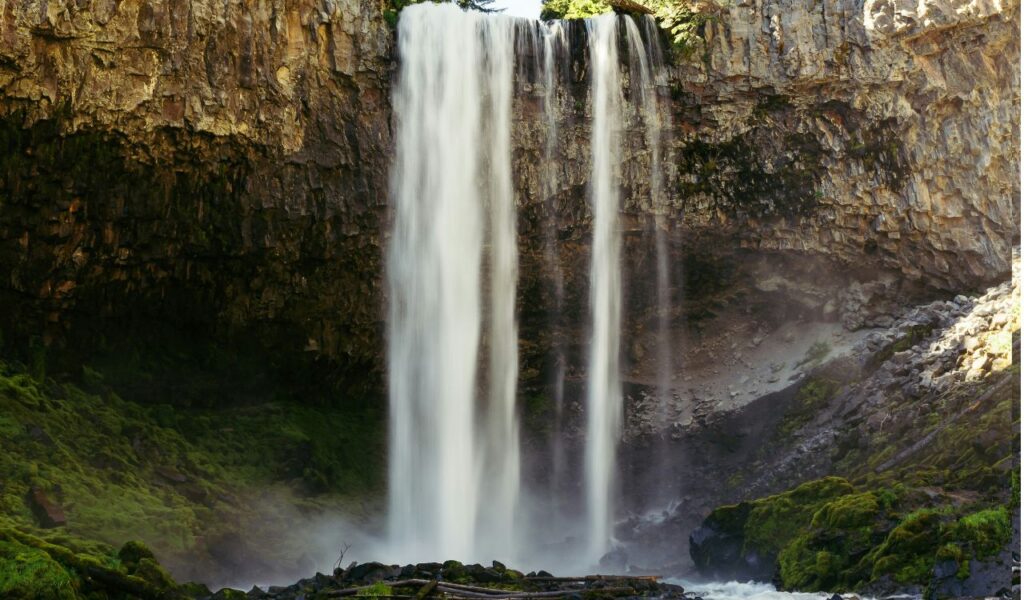 Tall twin streams of Soco Falls cascade from a rugged cliff into a misty pool below, surrounded by moss-covered rocks and lush greenery.