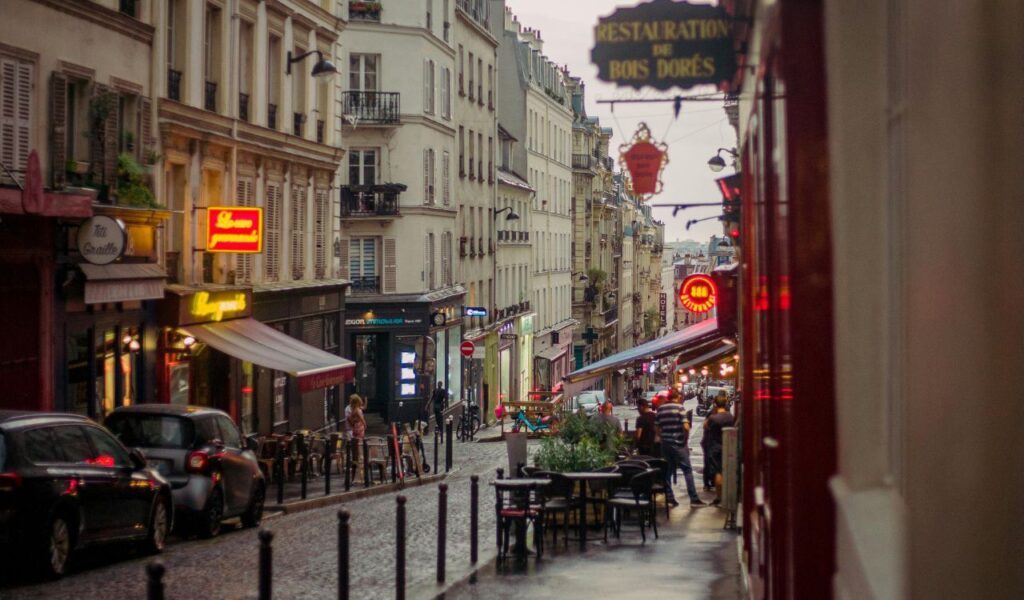A charming European street lined with cafes, shops, and neon signs, with outdoor seating and pedestrians walking on a rainy evening