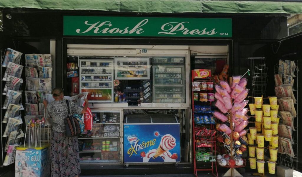 A small kiosk shop with a green sign labeled "Kiosk Press," displaying newspapers, snacks, cotton candy, and ice cream outside.