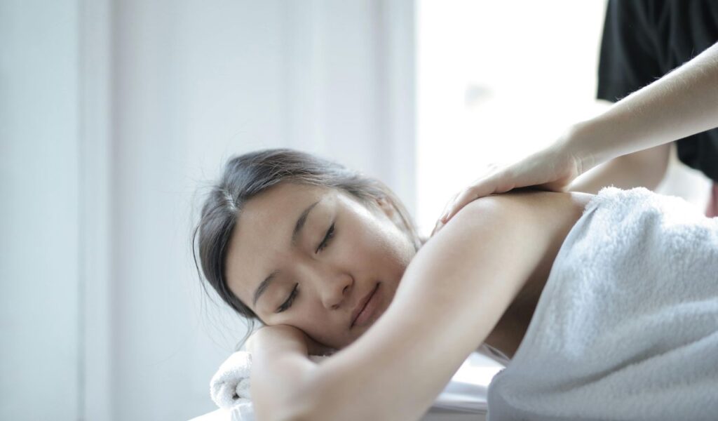 A woman lying on a massage table with a white towel, receiving a relaxing shoulder massage in a bright spa setting.
