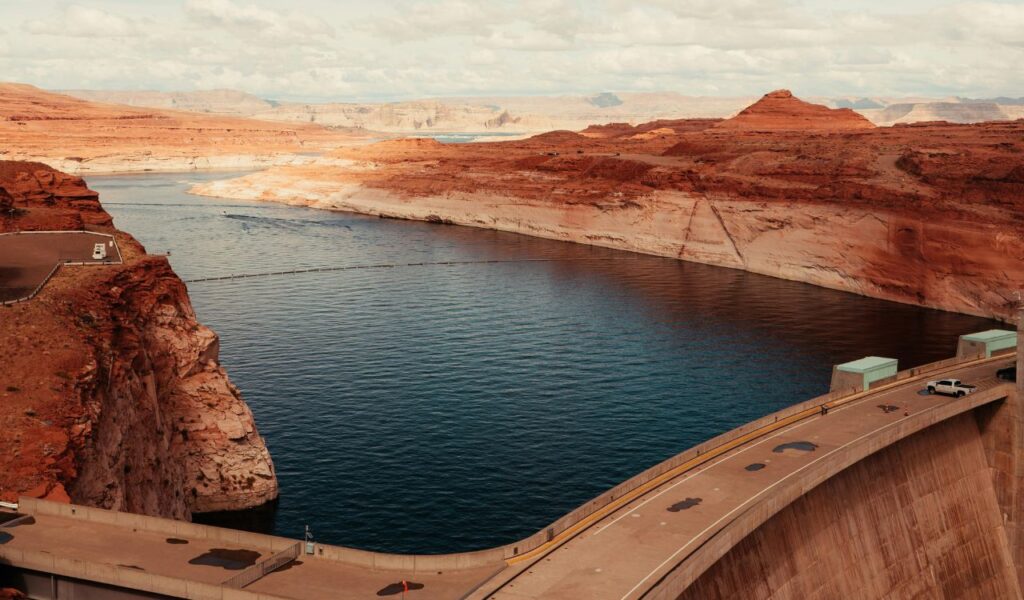 View of a dam with a curved road on top, overlooking a calm reservoir surrounded by red rocky cliffs and distant mesas.
