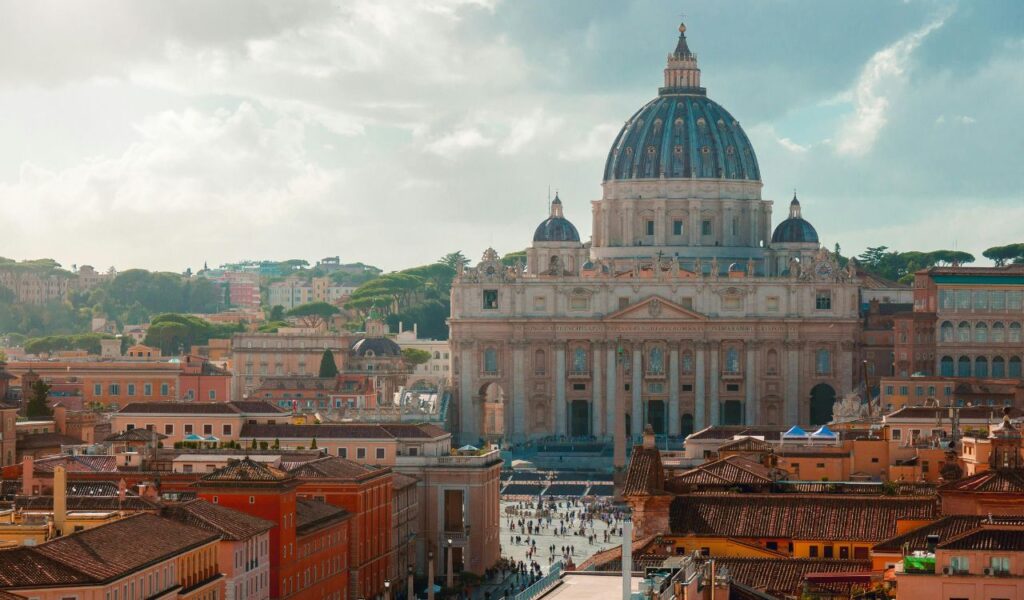 St. Peter’s Basilica in Vatican City, with its grand dome rising above the surrounding buildings and St. Peter’s Square filled with visitors, set against a backdrop of green hills and a partly cloudy sky.