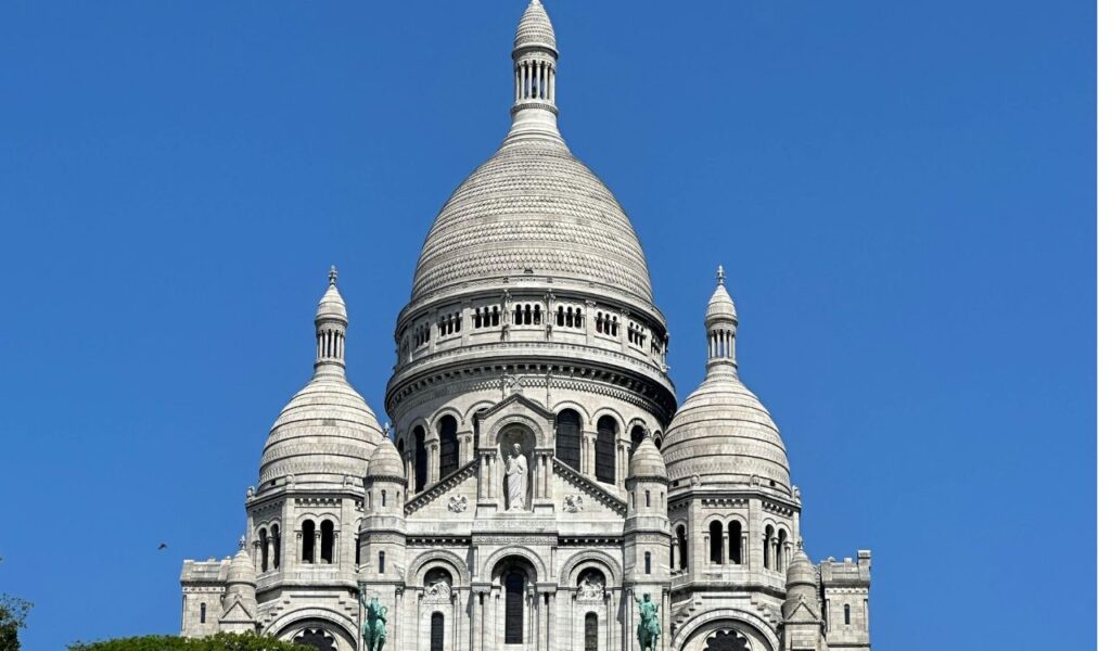 Sacré-Cœur Basilica in Paris, France, showcasing its striking white domes, intricate stone detailing, and statues, set against a vivid blue sky.