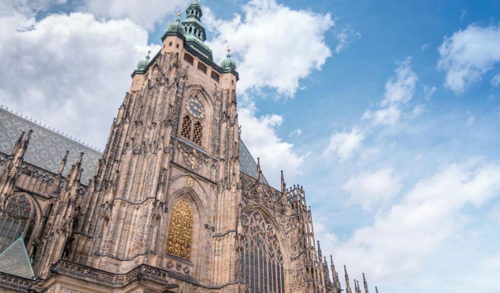 St. Vitus Cathedral in Prague, Czech Republic, featuring intricate Gothic architecture, a tall clock tower, ornate stained-glass windows, and detailed stone carvings under a partly cloudy sky.