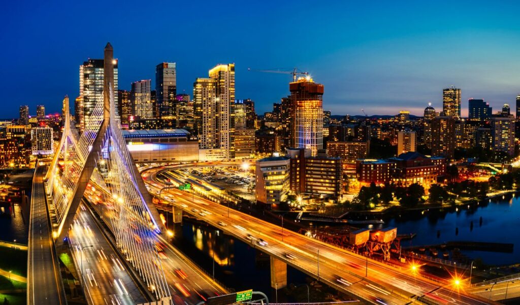 A vibrant nighttime cityscape featuring the Zakim Bridge and downtown Boston, Massachusetts, illuminated by bright lights and bustling traffic.
