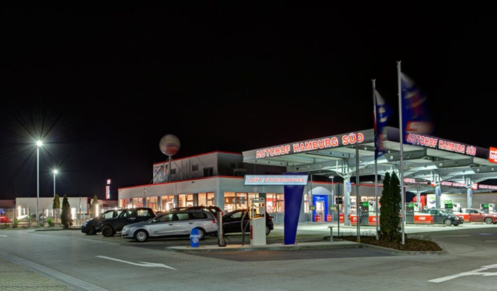 A brightly lit highway rest area at night with parked cars, fuel pumps, and a convenience store under a clear dark sky.