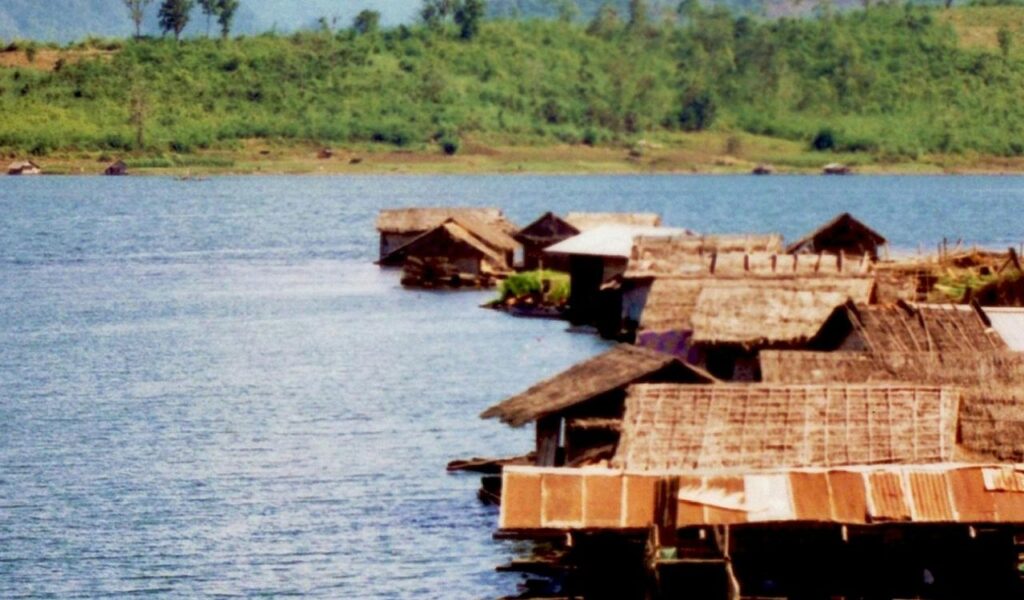 Traditional bamboo huts built on a tranquil lakeside in rural Thailand, surrounded by green hills and calm blue water under a clear sky.