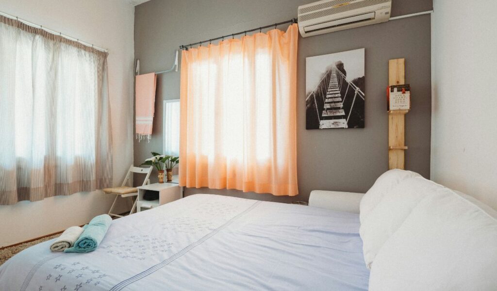 A neatly arranged bedroom with a made bed, folded towels, soft peach curtains, a wall-mounted air conditioner, and simple decor.