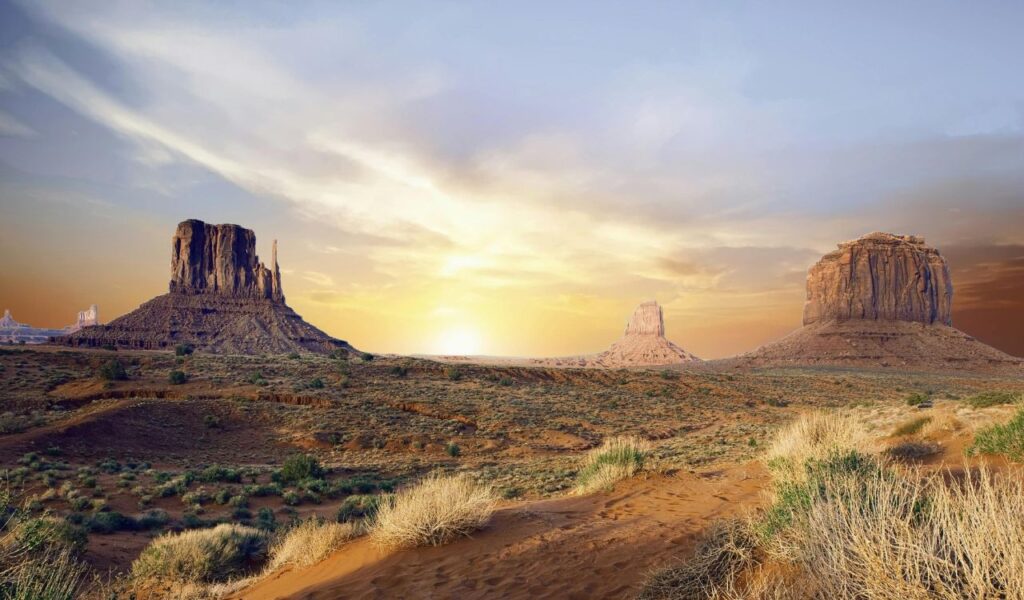Monument Valley at sunset with three towering sandstone buttes rising from the desert floor, surrounded by sparse vegetation and dramatic sky colors.