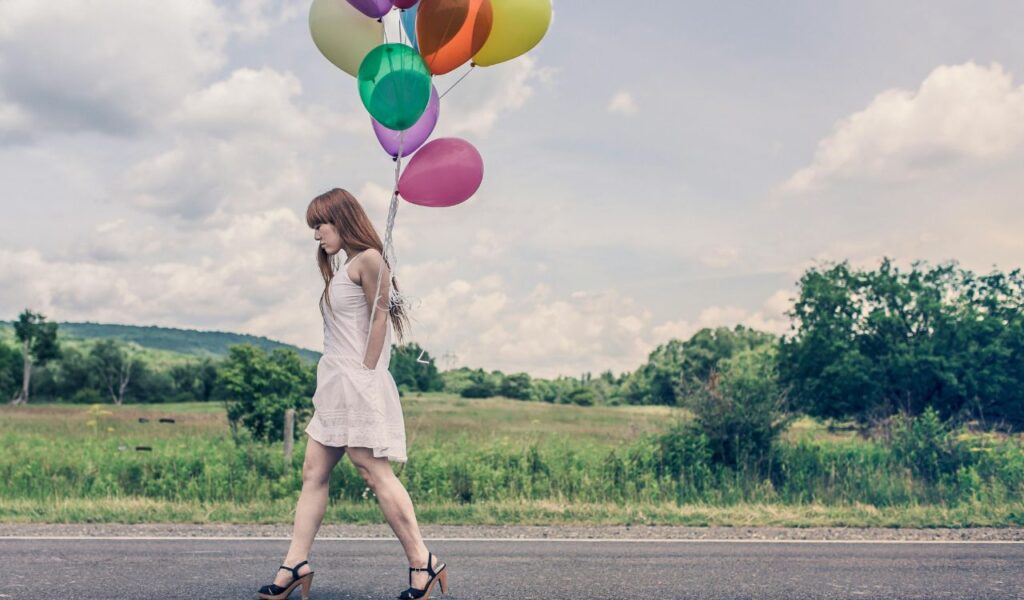 A young woman in a white dress and high heels walking along a rural road while holding a bunch of colorful balloons, with a grassy field and cloudy sky in the background.