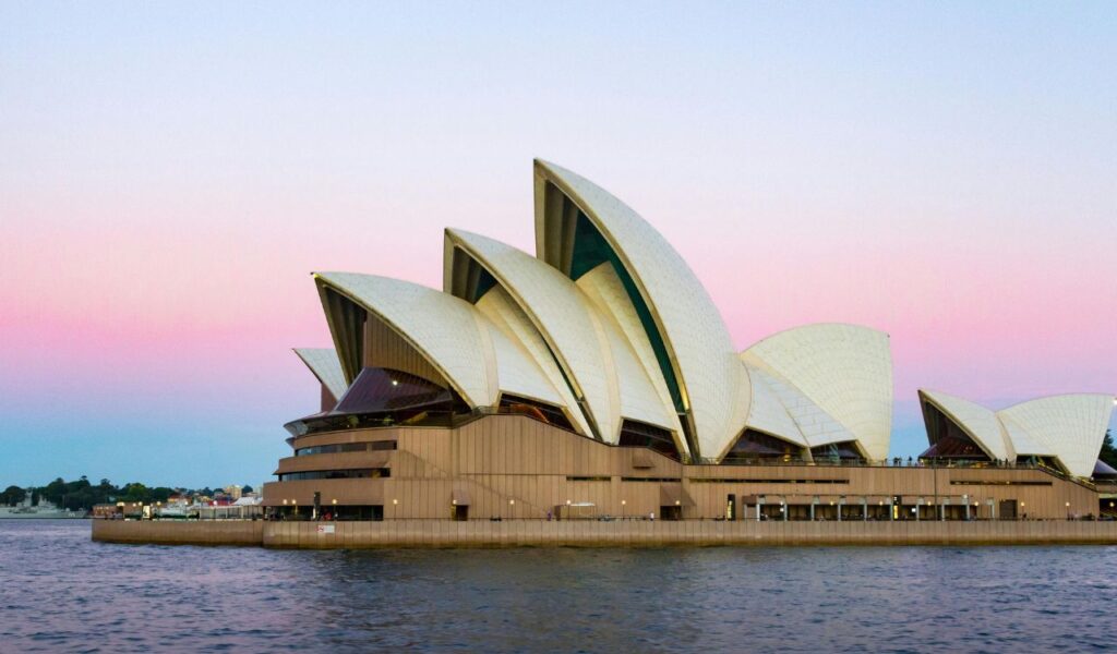 Sydney Opera House with its iconic sail-shaped design, viewed from the harbor at sunset with a pink and blue sky in the background.