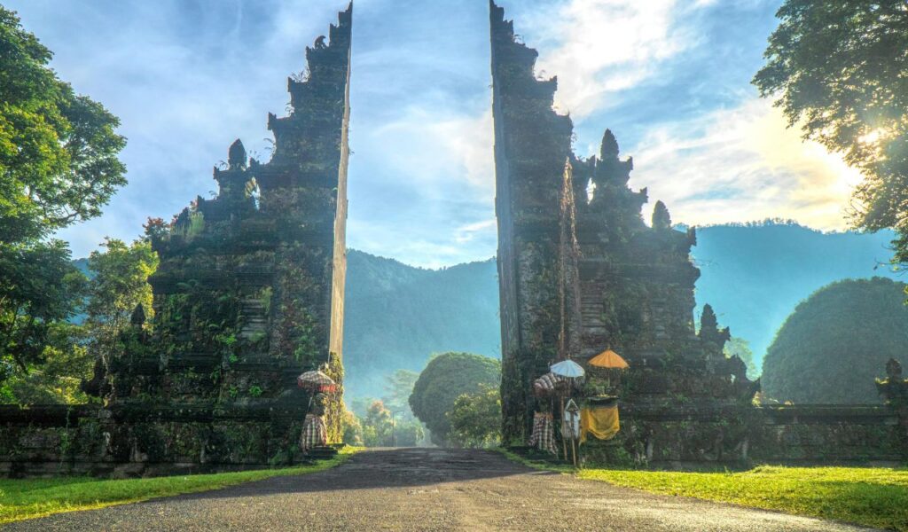 A traditional Balinese split gate surrounded by lush greenery and mountains under a bright, partly cloudy sky.