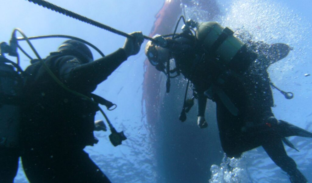 Two scuba divers hold onto a rope while ascending underwater, surrounded by bubbles, with sunlight filtering through the blue water above.