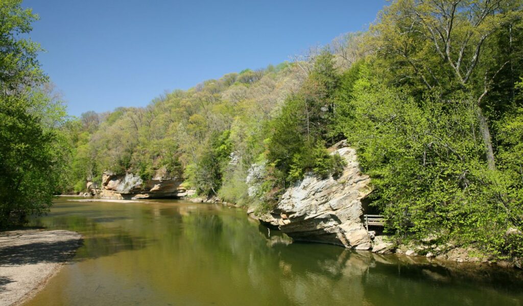 A calm river winds between rocky cliffs and lush green trees under a clear blue sky at Turkey Run State Park in Indiana.