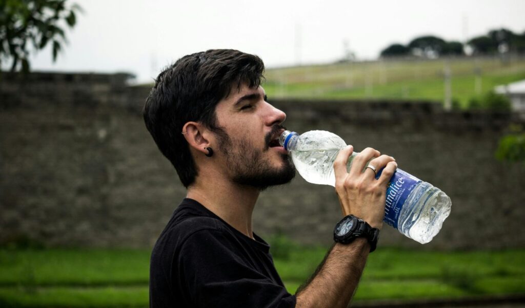 A man drinking from a water bottle outdoors on a hot day, with a blurred background of greenery and a stone wall.