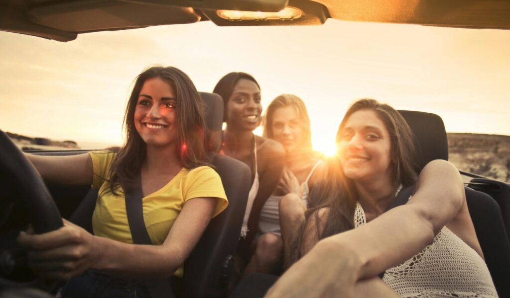 Four friends smiling and enjoying a car ride with the top down at sunset, creating a warm and carefree road trip vibe.