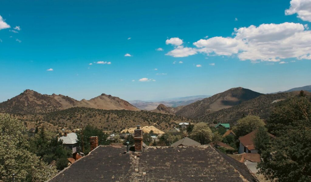 View of Virginia City, Nevada, with rustic rooftops, scattered houses, and surrounding desert mountains under a bright blue sky.