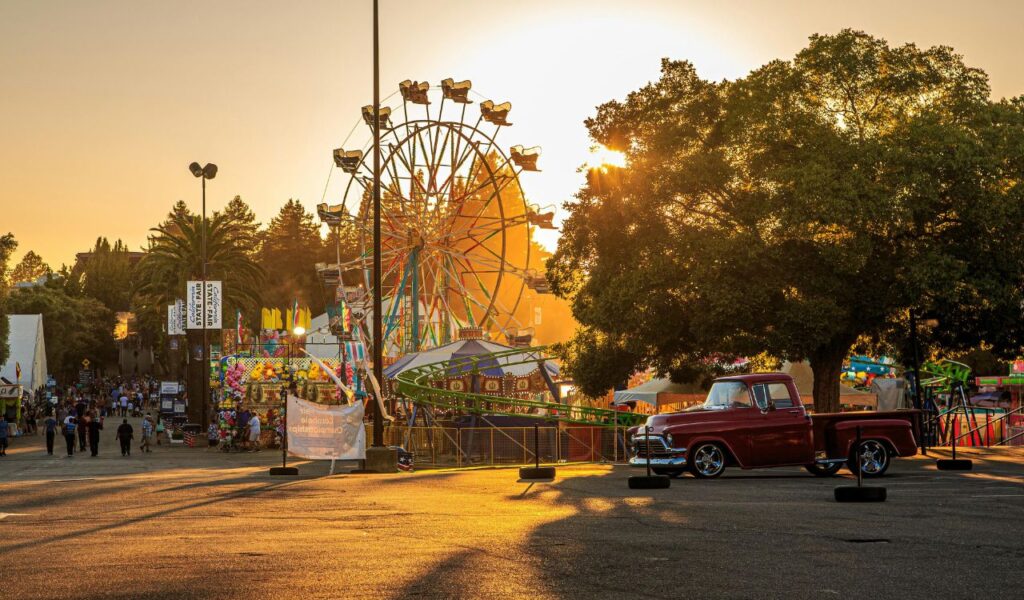 Sunset view of a state fair with a Ferris wheel, colorful rides, and game booths, alongside a classic red pickup truck parked under a large tree.