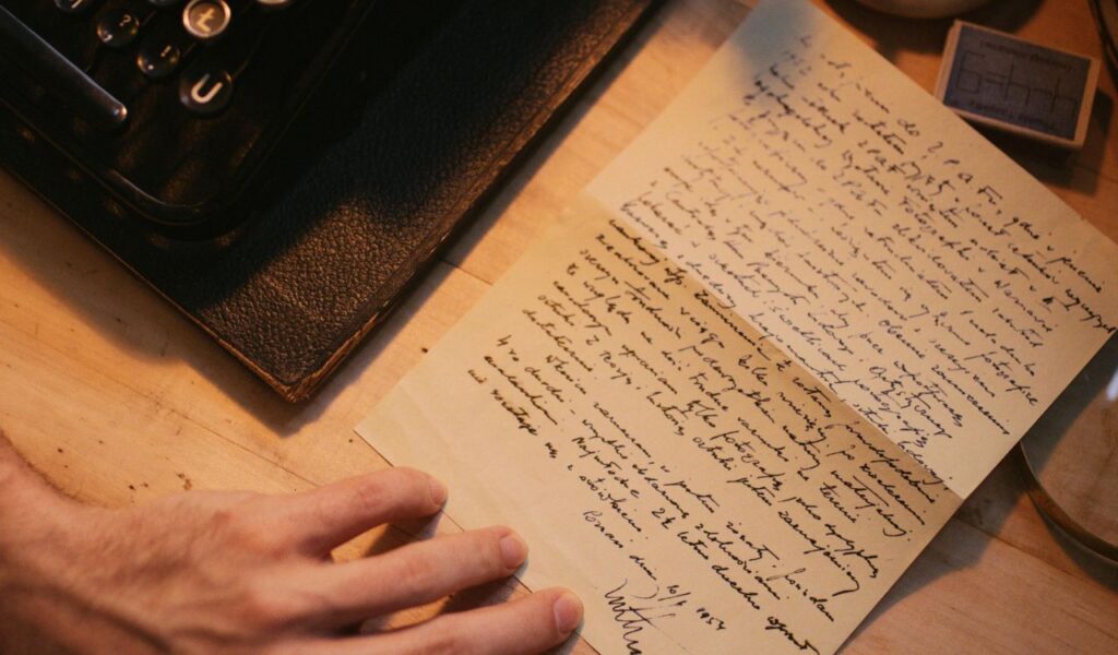 Close-up of a handwritten letter on a wooden desk next to a leather-bound book and a typewriter, with a person’s hand resting nearby.