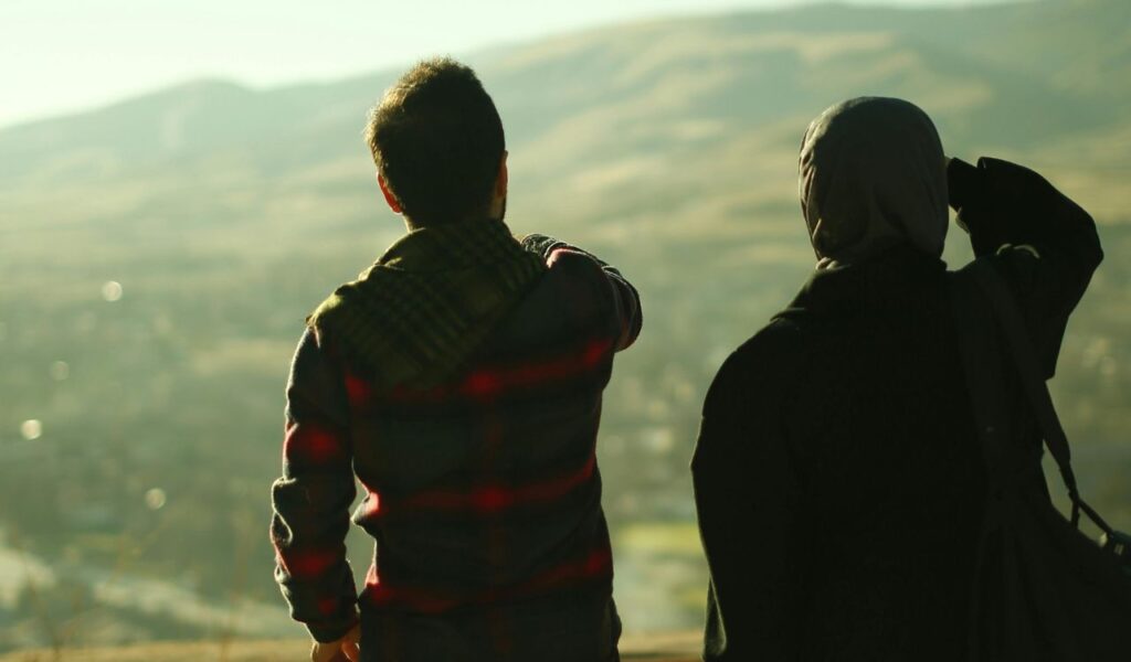 Two people standing outdoors, viewed from behind, watching a scenic landscape of rolling hills in the distance as one points toward the horizon.
