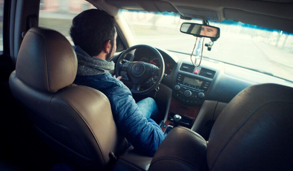 A man drives a car on an empty road, viewed from the backseat, with his reflection visible in the rearview mirror.