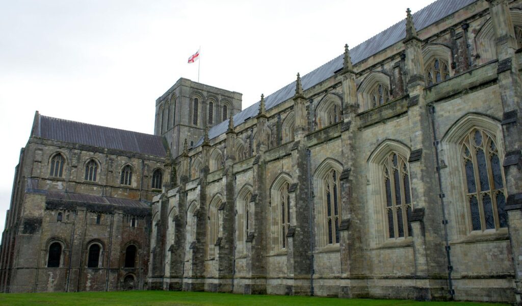  Exterior view of Winchester Cathedral with Gothic architecture, tall arched windows, and the flag of England flying above.