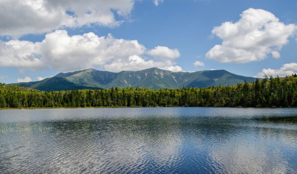 Calm lake in Wolfeboro, New Hampshire, reflecting green forests and the White Mountains under a bright blue sky with fluffy clouds