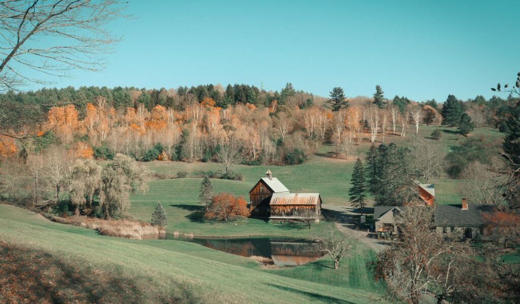 A peaceful autumn landscape in Woodstock, Vermont, featuring a rustic barn by a small pond, surrounded by rolling hills and colorful trees.