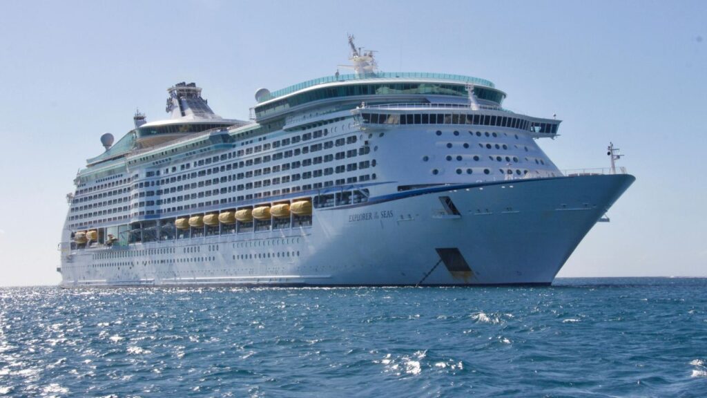 Large white cruise ship Explorer of the Seas anchored at sea on a sunny day, with yellow lifeboats and rows of balconies visible.