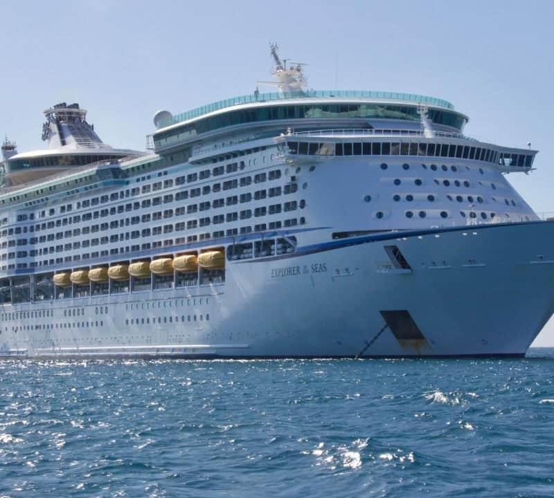 Large white cruise ship Explorer of the Seas anchored at sea on a sunny day, with yellow lifeboats and rows of balconies visible.