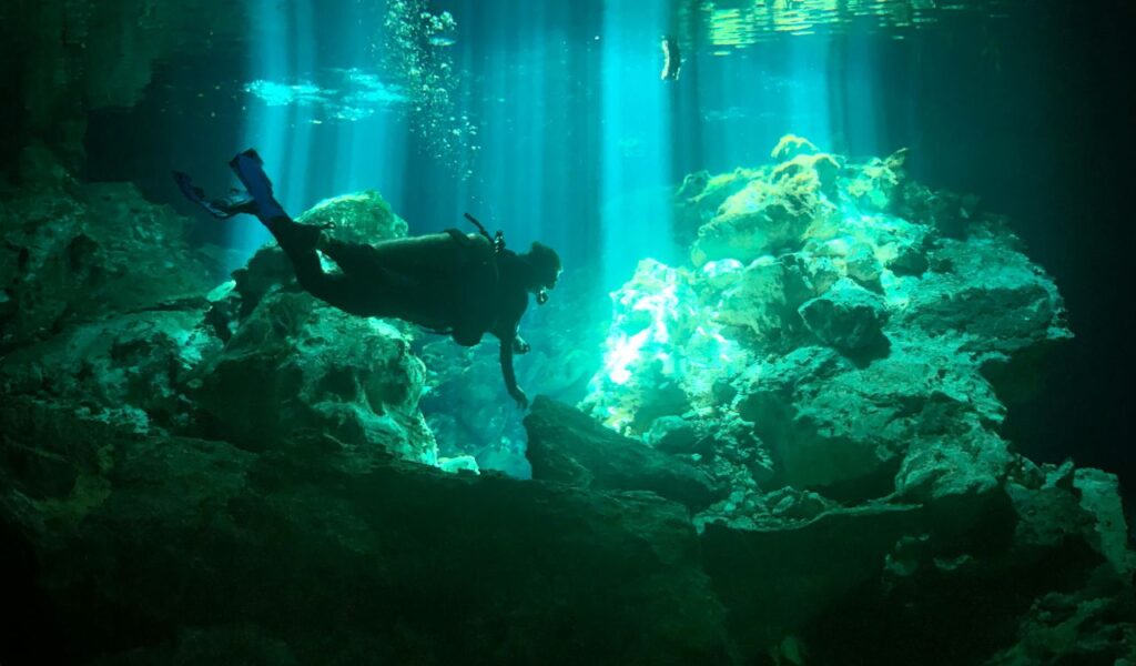A scuba diver explores an underwater cave illuminated by beams of light streaming from above, revealing rocky formations and clear water