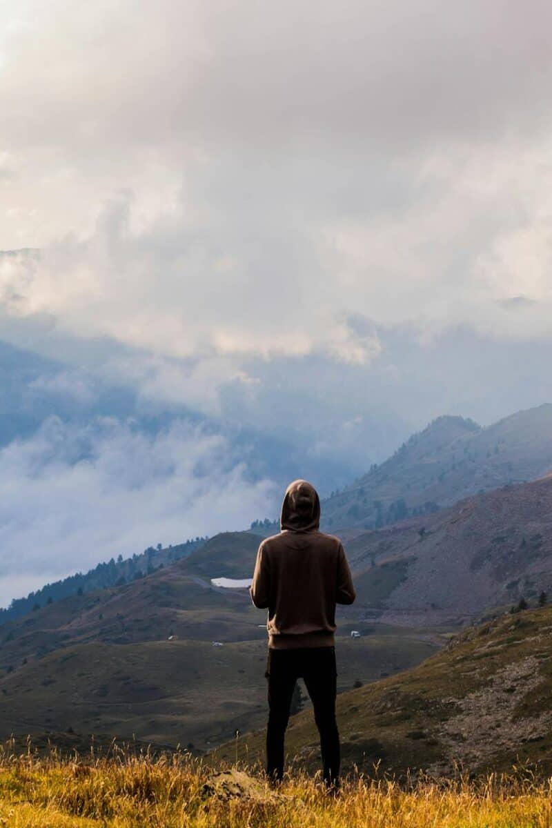 person wearing brown pullover and black jeans standing on mountain top with the scenic view